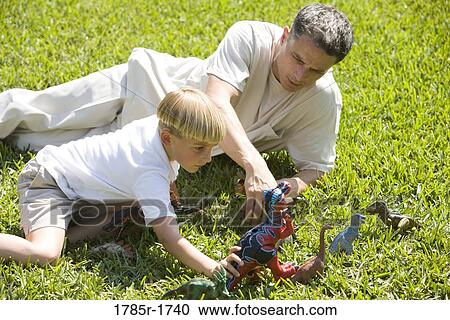 Stock Image - Father with son playing with toy dinosaurs on grassy lawn. Fotosearch