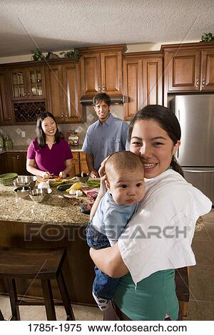 Girl holding baby while mom prepares dinner in background View Large Photo Image Stock Photography - Girl holding baby while mom prepares dinner in background. Fotosearch