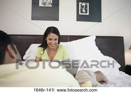 Stock Image - Mature woman sitting in front of a mature man with a laptop in the bed. Fotosearch