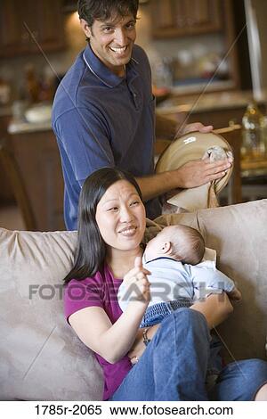 Stock Photography - Mother holding sleeping baby while her husband does household chores. Fotosearch