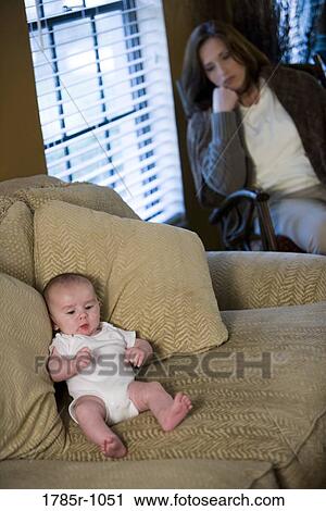 Stock Image - Mother suffering from postpartum depression, sitting by a window while the baby lies on a sofa. Fotosearch