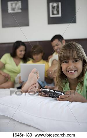 Portrait of a girl holding a handheld video game with her parents and brother looking at a laptop in the background View Large Photo Image Stock Photo - Portrait of a girl holding a handheld video game with her parents and brother looking at a laptop in the background. Fotosearch