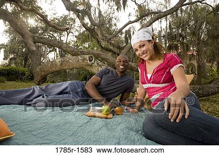 Stock Image - Portrait of a romantic young couple lying on a picnic blanket under oak trees. Fotosearch