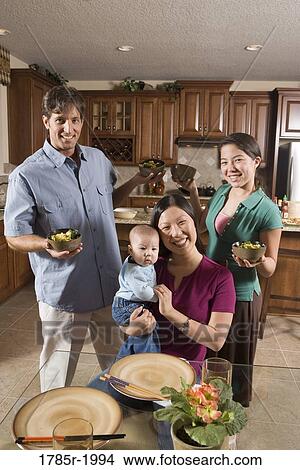 Picture - Portrait of an Asian-Caucasian family holding up bowls of food at a dining table. Fotosearch
