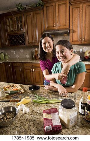 Stock Photography - Portrait of an Asian mother embracing her Asian-Caucasian daughter in the kitchen. Fotosearch