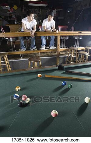 Stock Photograph - Portrait of two men in jeans and t-shirts sitting in front of billiards table. Fotosearch