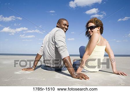 Rear view of a romantic young couple sitting on beach, turning around to look at viewer View Large Photo Image Stock Image - Rear view of a romantic young couple sitting on beach, turning around to look at viewer. Fotosearch