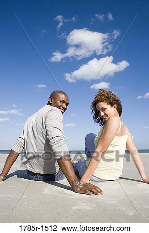 Rear view of a romantic young couple sitting on beach, turning around to look at viewer View Large Photo Image Stock Image - Rear view of a romantic young couple sitting on beach, turning around to look at viewer. Fotosearch
