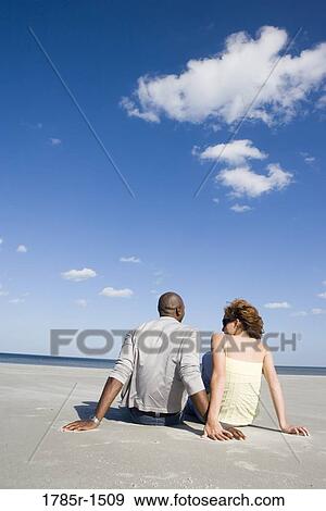 Stock Photo - Rear view of a romantic young couple sitting on beach. Fotosearch
