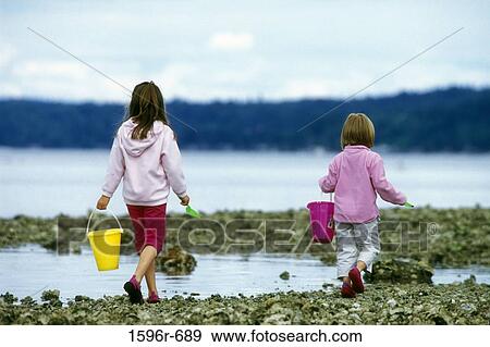 Stock Photo - Rear view of two girls carrying buckets and walking on the coast. Fotosearch