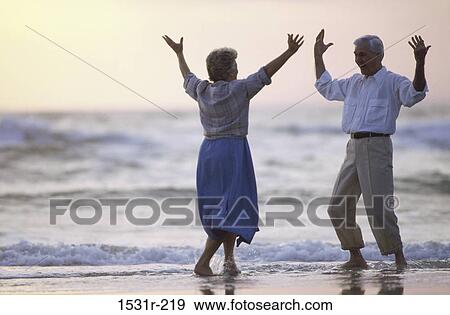Stock Photo - Senior couple standing on the beach with their arms raised. Fotosearch