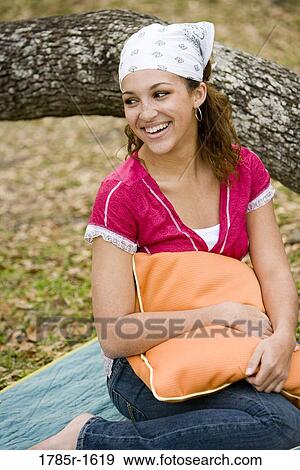 Smiling young woman holding a pillow sitting under a tree on a blanket View Large Photo Image Stock Photo - Smiling young woman holding a pillow sitting under a tree on a blanket. Fotosearch