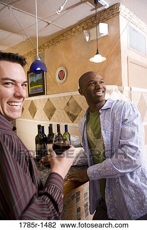 Stock Image - Two smiling young men standing by bar with wine bottles. Fotosearch