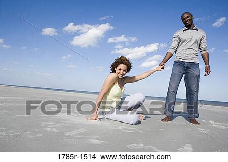 View of a smiling young woman sitting on beach, holding hands with standing man View Large Photo Image Picture - View of a smiling young woman sitting on beach, holding hands with standing man. Fotosearch