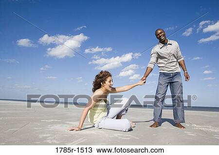 View of a smiling young woman sitting on beach, holding hands with standing man View Large Photo Image Stock Image - View of a smiling young woman sitting on beach, holding hands with standing man. Fotosearch