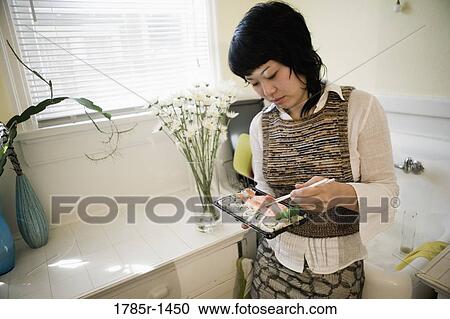 Stock Image - Young Asian woman eating sushi in kitchen. Fotosearch
