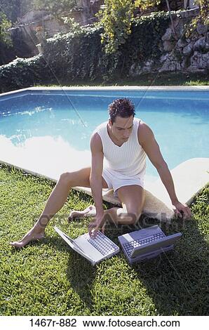 Stock Image - Young man sitting beside a swimming pool using a laptop. Fotosearch