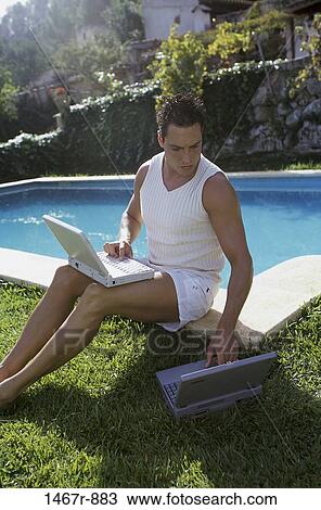 Stock Image - Young man sitting beside a swimming pool using a laptop. Fotosearch
