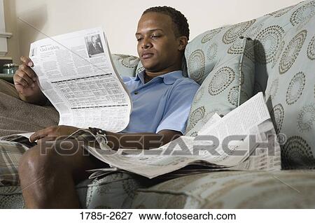 Stock Photo - Young man sitting on sofa and reading newspaper. Fotosearch