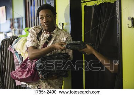 Stock Image - Young man standing in a boutique holding jeans outside fitting room. Fotosearch