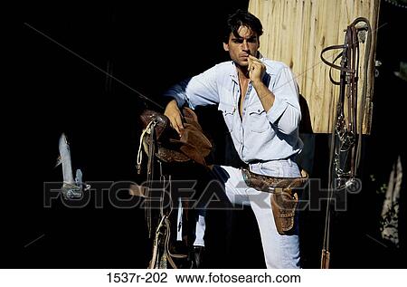 Stock Image - Young man standing near a saddle wearing a holster. Fotosearch