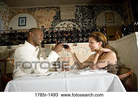 Stock Image - Young multi-ethnic couple at a restaurant table toasting with wine glasses. Fotosearch