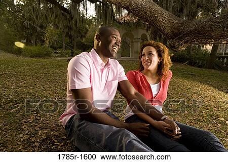Young multi-ethnic couple sitting on grass amidst oak trees at dusk View Large Photo Image Stock Image - Young multi-ethnic couple sitting on grass amidst oak trees at dusk. Fotosearch
