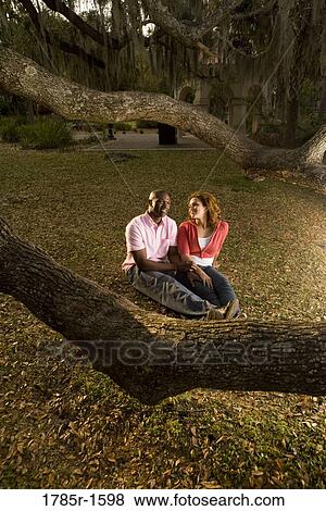 Young multi-ethnic couple sitting on grass amidst oak trees at dusk View Large Photo Image Stock Photo - Young multi-ethnic couple sitting on grass amidst oak trees at dusk. Fotosearch