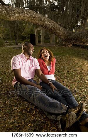 Young multi-ethnic couple sitting on grass amidst oak trees at dusk View Large Photo Image Stock Photo - Young multi-ethnic couple sitting on grass amidst oak trees at dusk. Fotosearch