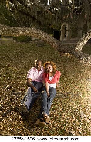 Young multi-ethnic couple sitting on grass amidst oak trees at dusk View Large Photo Image Stock Photograph - Young multi-ethnic couple sitting on grass amidst oak trees at dusk. Fotosearch
