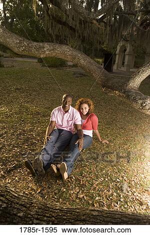 Stock Photography - Young multi-ethnic couple sitting on grass amidst oak trees at dusk. Fotosearch