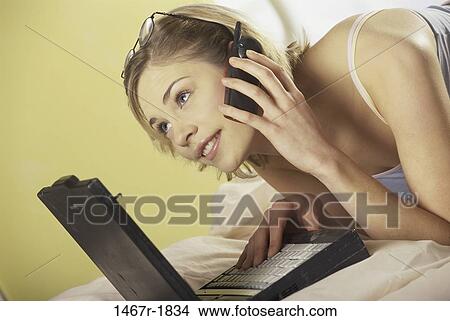 Young woman lying down in front of a laptop talking on a mobile phone View Large Photo Image Picture - Young woman lying down in front of a laptop talking on a mobile phone. Fotosearch