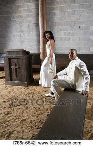Stock Image - A bride standing next to a groom seated on a bench, all in white. Fotosearch