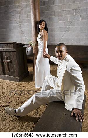 Stock Image - A bride standing next to a groom seated on a bench, all in white. Fotosearch