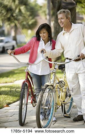Picture - A mature inter-racial couple walking with bicycles in their neighborhood. Fotosearch