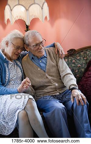 Stock Image - Affectionate enior couple smiling while sitting on sofa in living room. Fotosearch