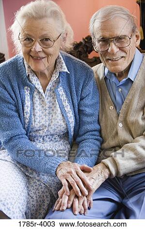 Stock Image - Affectionate enior couple smiling while sitting on sofa in living room. Fotosearch
