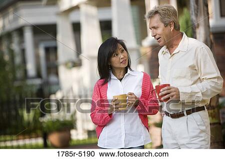 Stock Image - Affectionate mature inter-racial couple holding coffee mugs in front of their house. Fotosearch