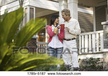 Stock Photo - Affectionate mature inter-racial couple holding coffee mugs in front of their house. Fotosearch