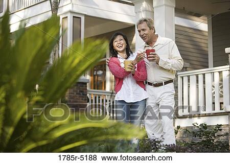 Stock Photo - Affectionate mature inter-racial couple holding coffee mugs in front of their house. Fotosearch