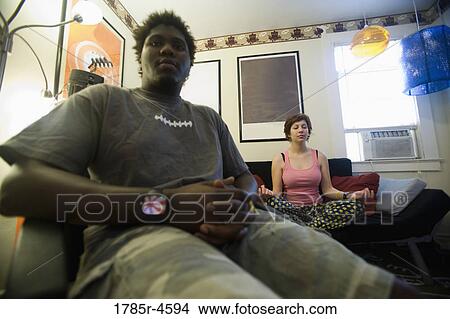 African man sitting in living room while woman meditates in background View Large Photo Image Picture - African man sitting in living room while woman meditates in background. Fotosearch