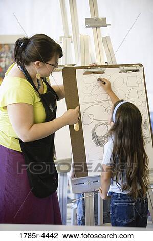 Stock Image - An art teacher showing young girl how to draw on an easel in art class. Fotosearch