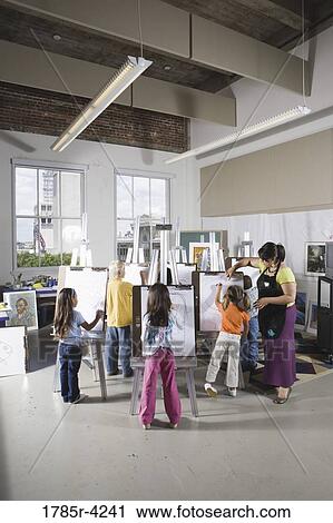 Stock Image - An art teacher with her students drawing on easels in art class. Fotosearch