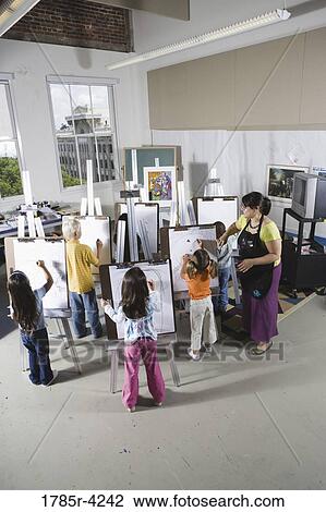 Stock Image - An art teacher with her students drawing on easels in art class. Fotosearch