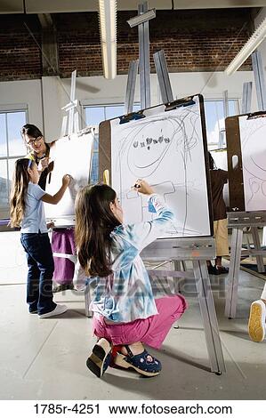 Stock Image - An art teacher with her students drawing on easels in art class. Fotosearch