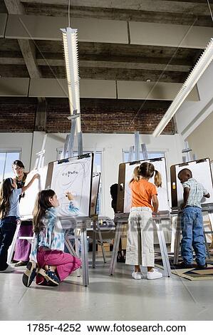 Stock Image - An art teacher with her students drawing on easels in art class. Fotosearch