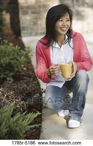 Stock Photograph - Asian woman holding coffee mug crouching on sidewalk. Fotosearch