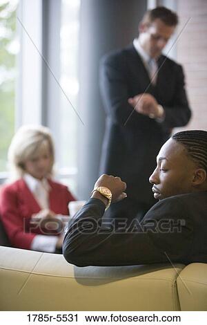 Stock Image - Business people sitting and waiting in an office lobby, looking at their watches. Fotosearch