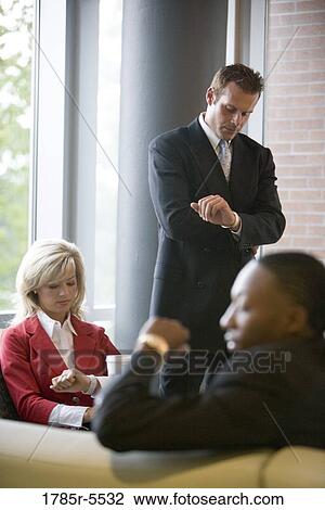 Stock Image - Business people sitting and waiting in an office lobby, looking at their watches. Fotosearch