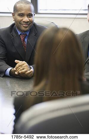 Stock Image - Businessman seated at conference table watching and listening to his colleague. Fotosearch